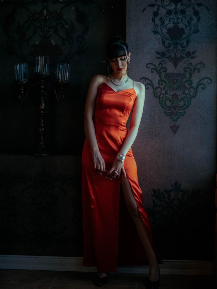 Woman in a sleek red dress posing indoors against an ornate dark wall, creating a moody ambiance.